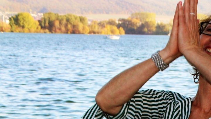 Eine Person praktiziert Gesichts-Yoga am Ufer eines Sees, umgeben von B&auml;umen und sanften H&uuml;geln im Hintergrund.