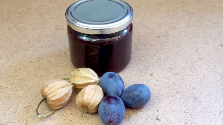 Ein Glas Pflaumenmarmelade steht neben frischen Physalis und Pflaumen auf einer hellen Oberfl&auml;che.