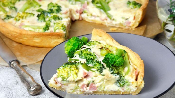 Ein Gem&uuml;sekuchen mit Brokkoli und Schinken steht auf einem Tisch, w&auml;hrend ein St&uuml;ck auf einem grauen Teller serviert wird.