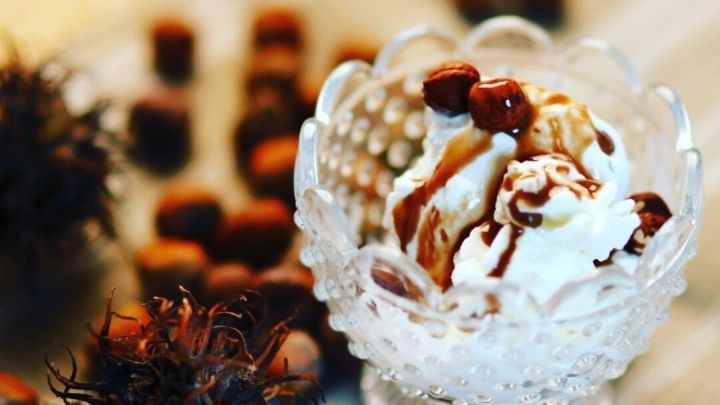 Ein h&uuml;bsches Dessert mit Sahne und Schokoladensauce steht in einem Kristallglas, umgeben von Haseln&uuml;ssen und einer Tischdecke.