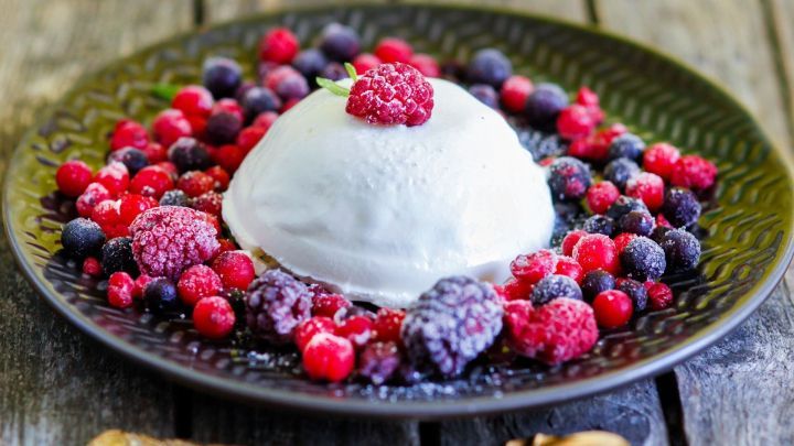 Eine Joghurt-Bombe sitzt auf einem dekorativen Teller, umgeben von frischen Beeren auf einem rustikalen Holztisch.