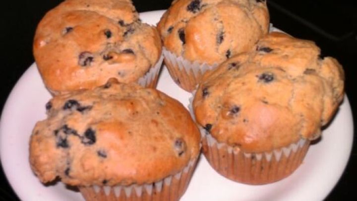 Vier Blaubeer-Muffins liegen auf einem wei&szlig;en Teller. Saftige, dunkle Blaubeeren sind in den lockeren Teig gebacken.