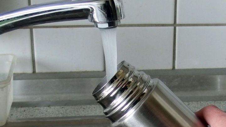 Eine handgehaltene Thermoskanne wird mit Wasser aus einem Wasserhahn gef&uuml;llt, w&auml;hrend die K&uuml;chenoberfl&auml;che im Hintergrund sichtbar ist.