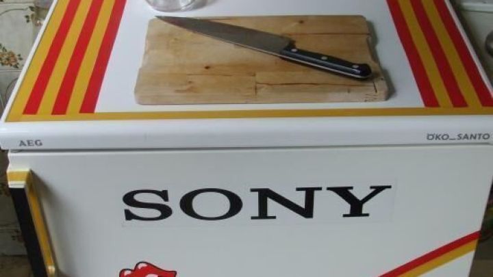 Ein verzierter K&uuml;hlschrank hat bunte Streifen und Sticker, steht in einer K&uuml;che mit Schneidebrett, Messer und Utensilien darauf. Text: "SONY".