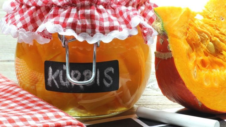 Ein Glas mit eingelegtem K&uuml;rbis steht auf einem Tisch, umgeben von einem halben K&uuml;rbis und einem karierten Tuch.