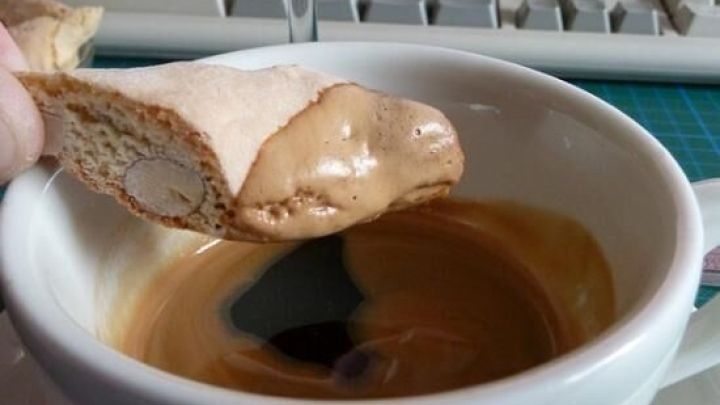 Eine Cantuccini wird in eine Tasse Kaffee getunkt, mit einem Weinglas und einer Computertastatur im Hintergrund. Text auf der Tasse: "bristat".