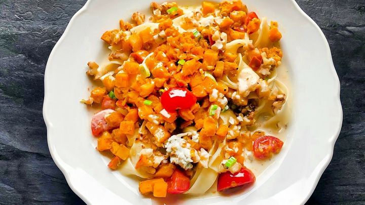 Ein Teller mit &uuml;berbackenem K&uuml;rbis, frischen Tomaten und Nudel-Rahmso&szlig;e ruht auf einem dunklen Untergrund.