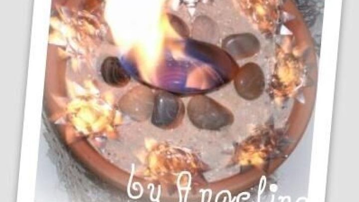 Ein brennendes Tischfeuer, umgeben von dekorativen Steinen, in einem Topf auf Sand. Text am Bildrand: "by Angelino".