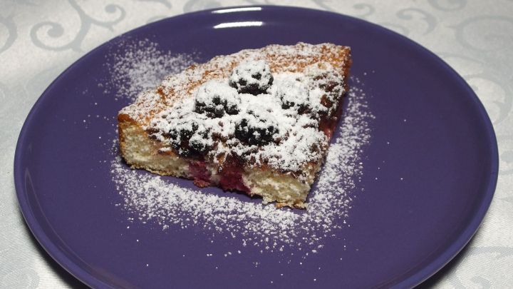 Ein St&uuml;ck Kuchens mit Kirschen liegt auf einem lila Teller, bestreut mit Puderzucker, auf einem wei&szlig;en Tisch.