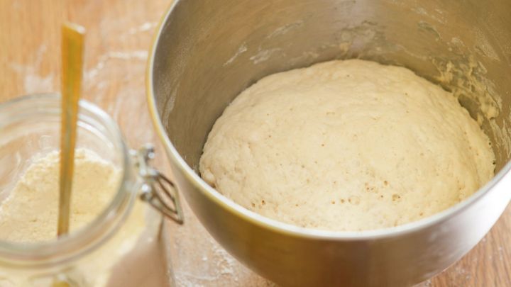 Ein R&uuml;hrsch&uuml;ssel mit Hefeteig steht auf einem Tisch, w&auml;hrend der Teig in kaltem Wasser aufgeht. Neben der Sch&uuml;ssel ist ein Glas mit Mehl.