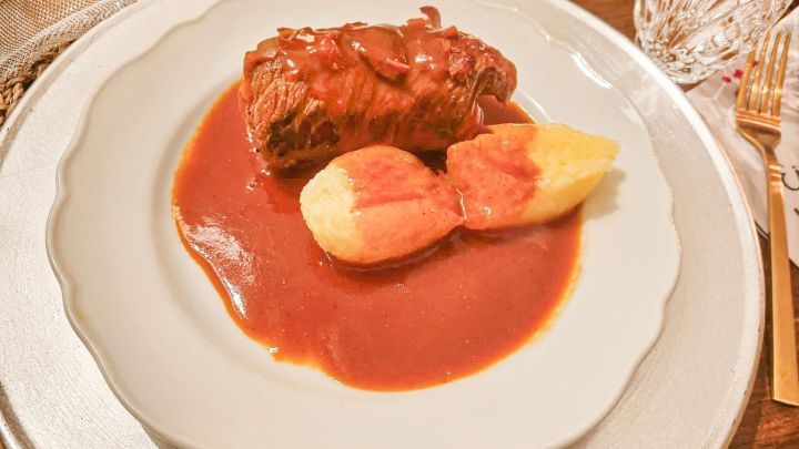 Eine gef&uuml;llte Rinderroulade liegt auf einem Teller, umgeben von einer w&uuml;rzigen Sauce und Beilagen wie Kartoffeln.