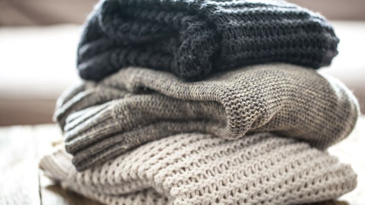 Gestapelt liegen drei Kratzende Wollpullover auf einem Holztablett, umgeben von einem hellen, unscharfen Hintergrund.