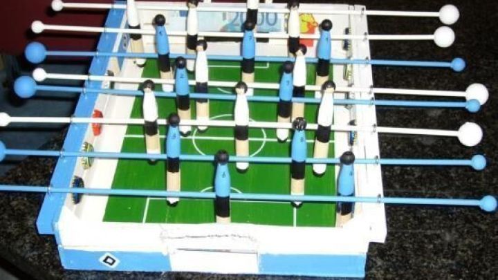 Tischfu&szlig;ballspiel mit Spielfiguren auf gr&uuml;nem Feld; gebaut auf schwarzem Tisch. Holzstangen halten blau-wei&szlig;e Spielerfiguren.