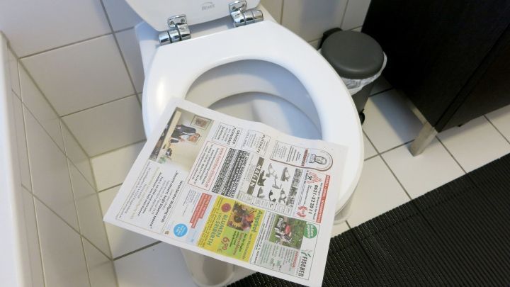 Klobrille mit ge&ouml;ffnetem Deckel, eine Zeitung daraufliegend, in einem gefliesten Badezimmer mit M&uuml;lleimer und dunklem Teppich.