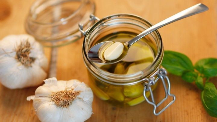 Ein Glas mit Knoblauch in &Ouml;l steht auf einem Holztisch, daneben liegen frische Knoblauchzehen und ein gr&uuml;nes Blatt.