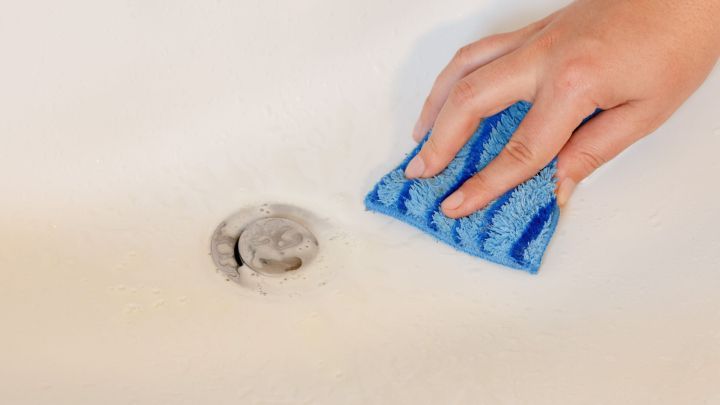 Eine Hand mit einem blauen Schwamm reinigt eine Badewanne neben einem Abfluss, um das Essigreiniger-Ergebnis zu verbessern.