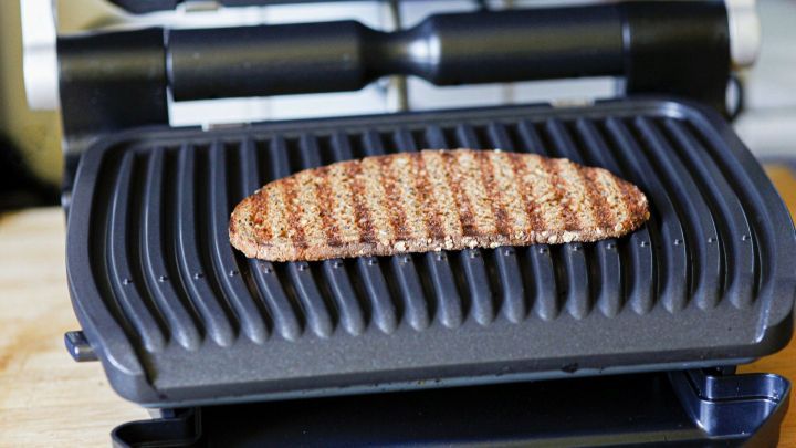 Ein Kontaktgrill grillt ein saftiges St&uuml;ck Fleisch, w&auml;hrend er auf einer Holzfl&auml;che in einer gem&uuml;tlichen K&uuml;che steht.
