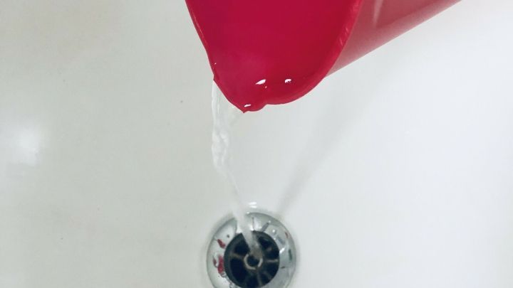 Ein roter Eimer kippt &uuml;ber eine Abfluss&ouml;ffnung, wobei Wasser hineingeht und das verstopfte Abflussproblem verbessert.