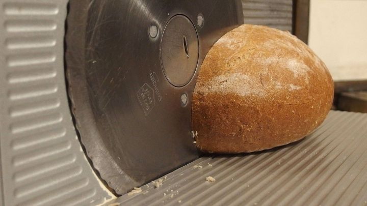 Eine Brotschneidemaschine schneidet ein Brot in der N&auml;he, w&auml;hrend Mehlkr&uuml;mel sich auf der Oberfl&auml;che sammeln.