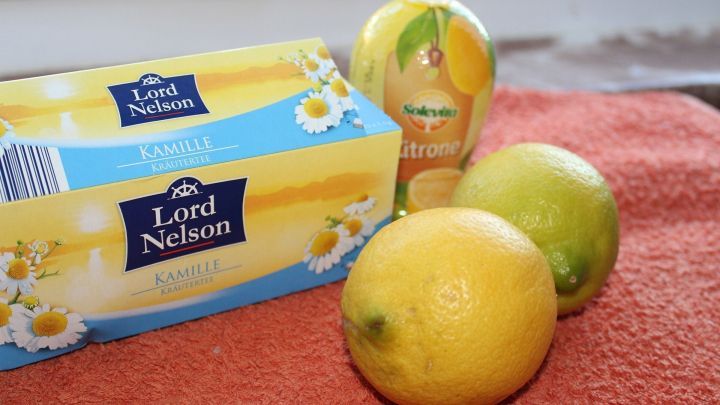 Zwei Zitronen und eine Flasche Zitronensaft liegen auf einem Handtuch neben einer Teeverpackung in einer hellen Umgebung.