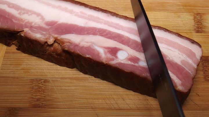 Ein dicker Streifen Speck liegt auf einem Holzbrett, w&auml;hrend ein scharfer Messer einen Schnitt ansetzt, um ihn zu teilen.