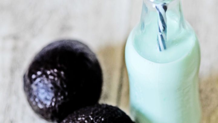 Ein gr&uuml;ner Avocado-Kokos-Vanille-Smoothie steht in einer Glasflasche mit Strohhalm neben zwei reifen Avocados auf einem Holztisch.