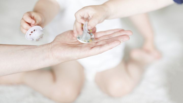 Zwei H&auml;nde halten Schnuller, w&auml;hrend ein Kind im wei&szlig;en Outfit auf einem weichen Teppich sitzt und zuschaut.