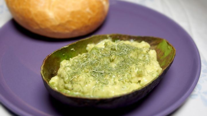 Ein gr&uuml;ner Avocado-Aufstrich liegt auf einem lila Teller neben einem frischen Br&ouml;tchen, bereit f&uuml;rs Fr&uuml;hst&uuml;ck.