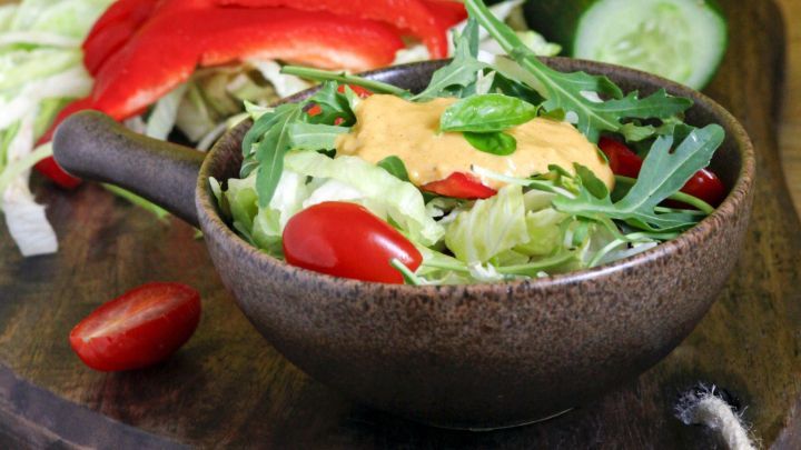 Bunte Salate in einer Sch&uuml;ssel mit Dressing, umgeben von frischen Zutaten wie Gurke und Paprika auf einem Holzbrett.