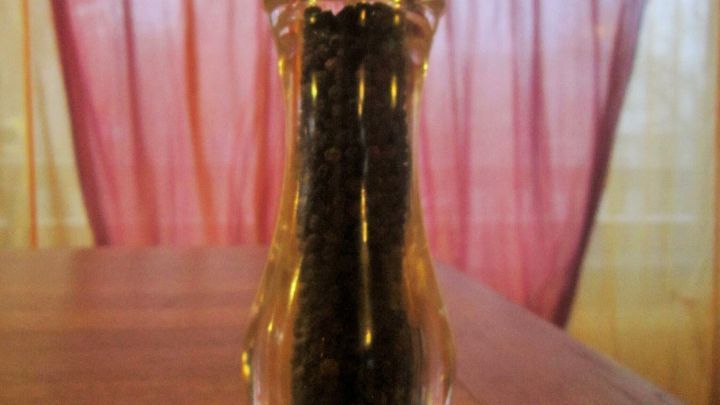 Eine transparente Pfefferm&uuml;hle steht auf einem Holztisch und zeigt ihre gef&uuml;llte Innenseite vor einem roten Vorhang.