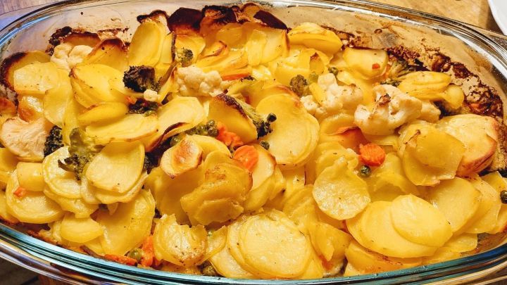 Ein frisch gebackenes Kartoffelgericht liegt in einer Glasform, umgeben von buntem Gem&uuml;se und goldbraunen Kartoffelscheiben.