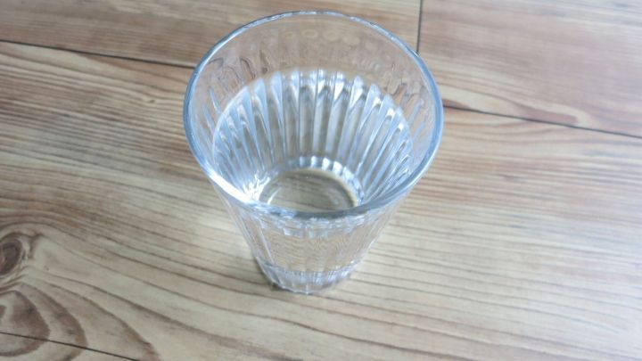 Ein transparentes Glas mit klarem Wasser steht auf einem Holzboden und reflektiert das Licht. Es symbolisiert Wasser als gesund und kalorienverbrennend.
