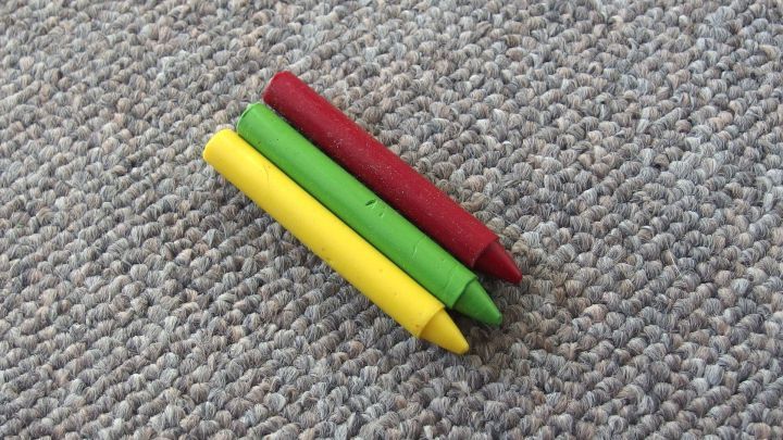 Drei Wachsfarben in Rot, Gr&uuml;n und Gelb liegen auf einem Teppich, bereit, beim Entfernen von Wachsfarbenflecken zu helfen.