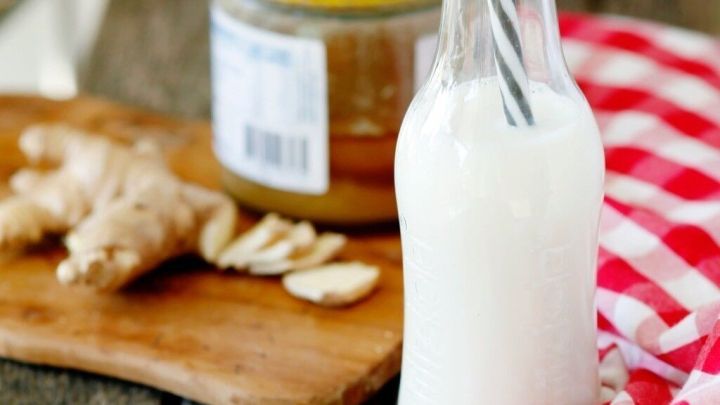 Eine Glasflasche mit einem Strohhalm steht auf einem Holzbrett neben Ingwer und Honig, umgeben von einem karierten Tuch.