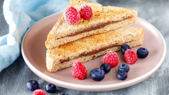Drei gegrillte Schoko-Sandwiches auf einem Teller, umgeben von frischen Himbeeren und Heidelbeeren, auf einem hellen Untergrund.