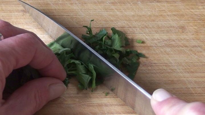 Eine Hand schneidet frisch gehackte Petersilie mit einem scharfen Messer auf einem Holzbrett.