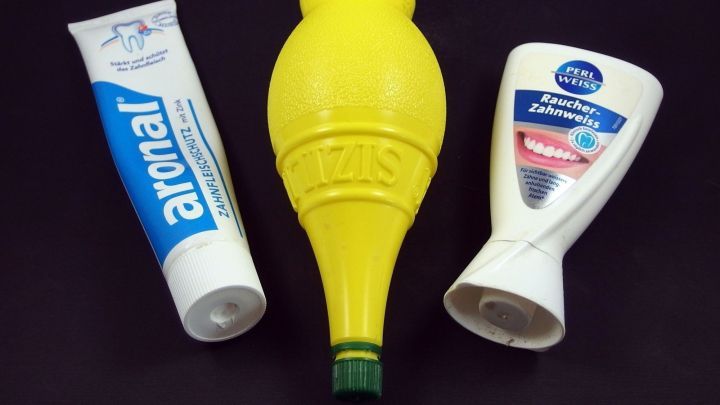 Eine Zahnpasta-Tube und eine Flasche in Zitronenform stehen neben einer zweiten Zahnpastatube auf einem dunklen Hintergrund.