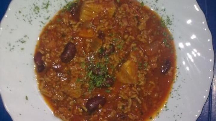Ein Teller mit scharfem Chilli con Carne, mit Fleisch und Bohnen, garniert mit gr&uuml;nen Kr&auml;utern auf einem blauen Tisch.