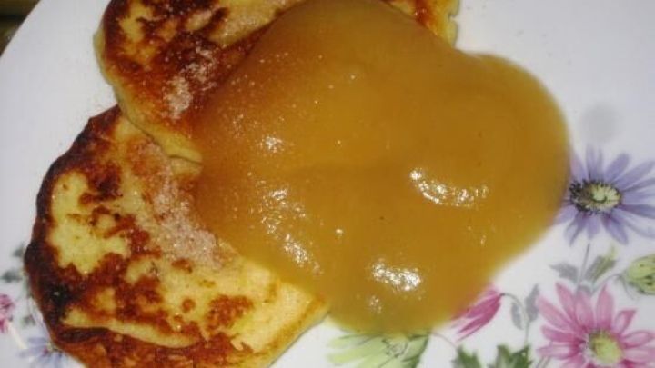 Zwei Quarkplinsen liegen auf einem Teller mit Blumenmuster, bedeckt mit einem gro&szlig;en Klecks Apfelmus.