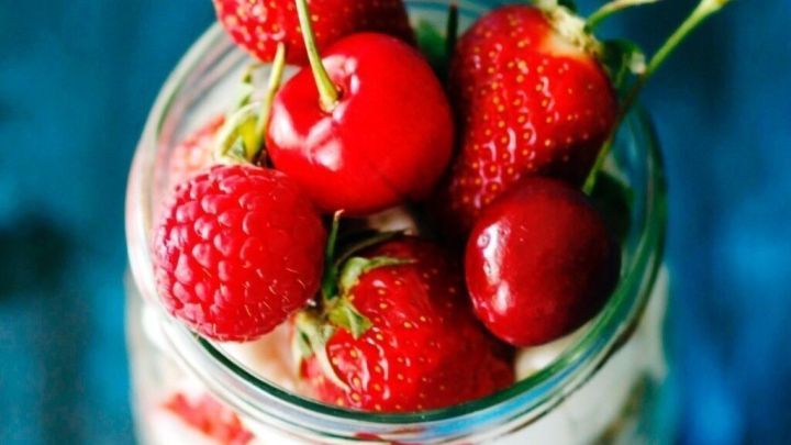 Ein glas Mini-Tiramisu ist gef&uuml;llt mit einer Schicht Sahne und frischen Himbeeren sowie Kirschen, auf einem blauen Untergrund.