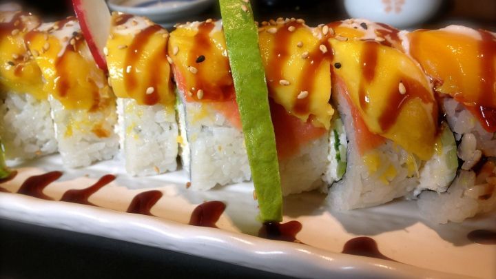 Ein farbenfrohes Sushi-Rollen-Arrangement liegt auf einem wei&szlig;en Teller, umgeben von dunklen Tischen und der K&uuml;che im Hintergrund.