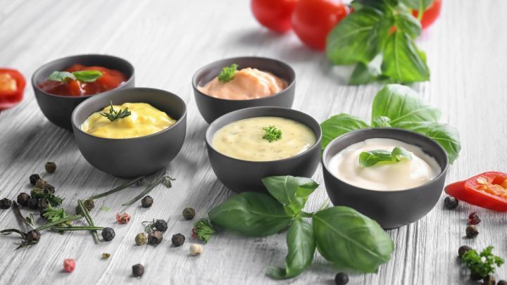 F&uuml;nf Schalen mit verschiedenen Grill- und Fondueso&szlig;en stehen auf einem Holztablett, umgeben von frischem Gem&uuml;se und Kr&auml;utern.