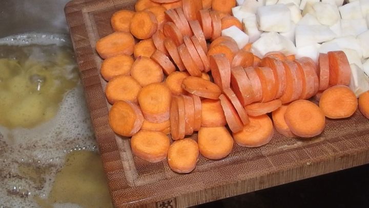 Auf einem Holzbrett liegen geschnittene M&ouml;hren und Kohlrabi, w&auml;hrend im Hintergrund eine Kartoffelsuppe kocht.