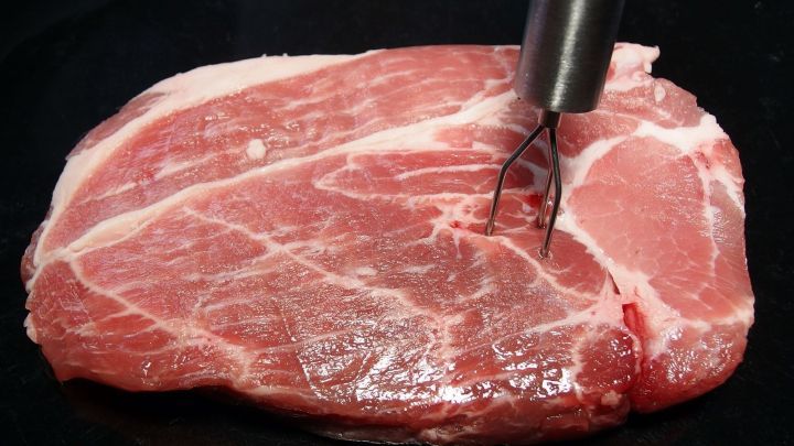 Eine Fleischzartmacher-Gabel wird in ein St&uuml;ck rohes Fleisch gesteckt, um es zarter zu machen, vor einem dunklen Hintergrund.