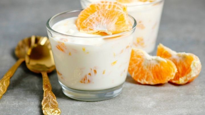 Zwei Teelichtgl&auml;ser beinhalten ein cremiges Dessert mit Mandarinenst&uuml;cken, umgeben von goldenen L&ouml;ffeln und Fruchtst&uuml;cken.