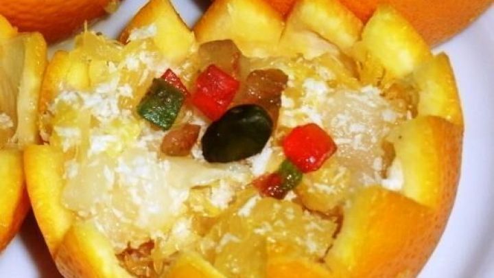 Ein Orangenk&ouml;rbchen steht auf einem Teller, gef&uuml;llt mit einem s&uuml;&szlig;en Pina-Colada-Dessert und bunten, kandierten Fr&uuml;chten.