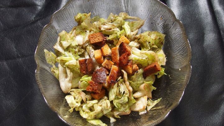 Ein Salat mit St&uuml;cken von altbackenen Br&ouml;tchen wird in einer Glas-Sch&uuml;ssel serviert. Der Salat liegt auf einem schwarzen Untergrund.
