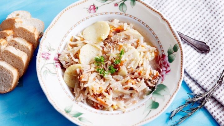 Eine Sch&uuml;ssel Wei&szlig;kohlsuppe mit Fleisch und Kr&auml;utern steht auf einem blauen Tisch, umgeben von Brot und einer Serviette.