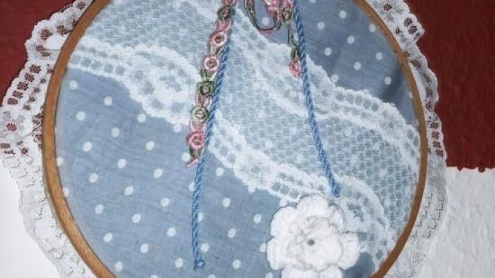 Ein runder, bestickter Stickrahmen mit blauer, gepunkteter Stoffunterlage h&auml;ngt an einer Wand und zeigt eine filigrane Spitzenborte.