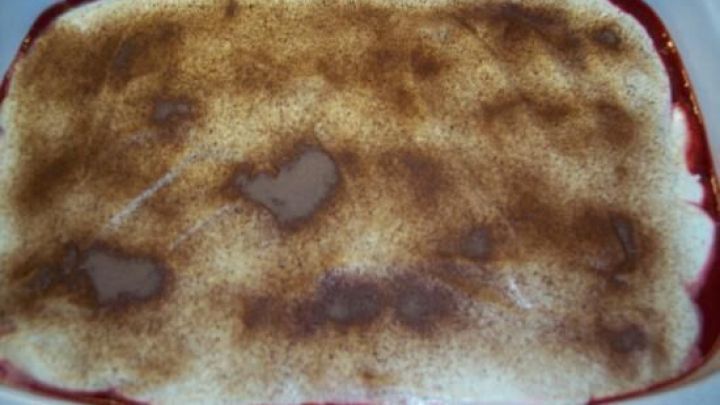 Ein Tiramisu mit einer goldbraunen Oberfl&auml;che liegt in einer wei&szlig;en Auflaufform, umgeben von fruchtig-roten Schichten.